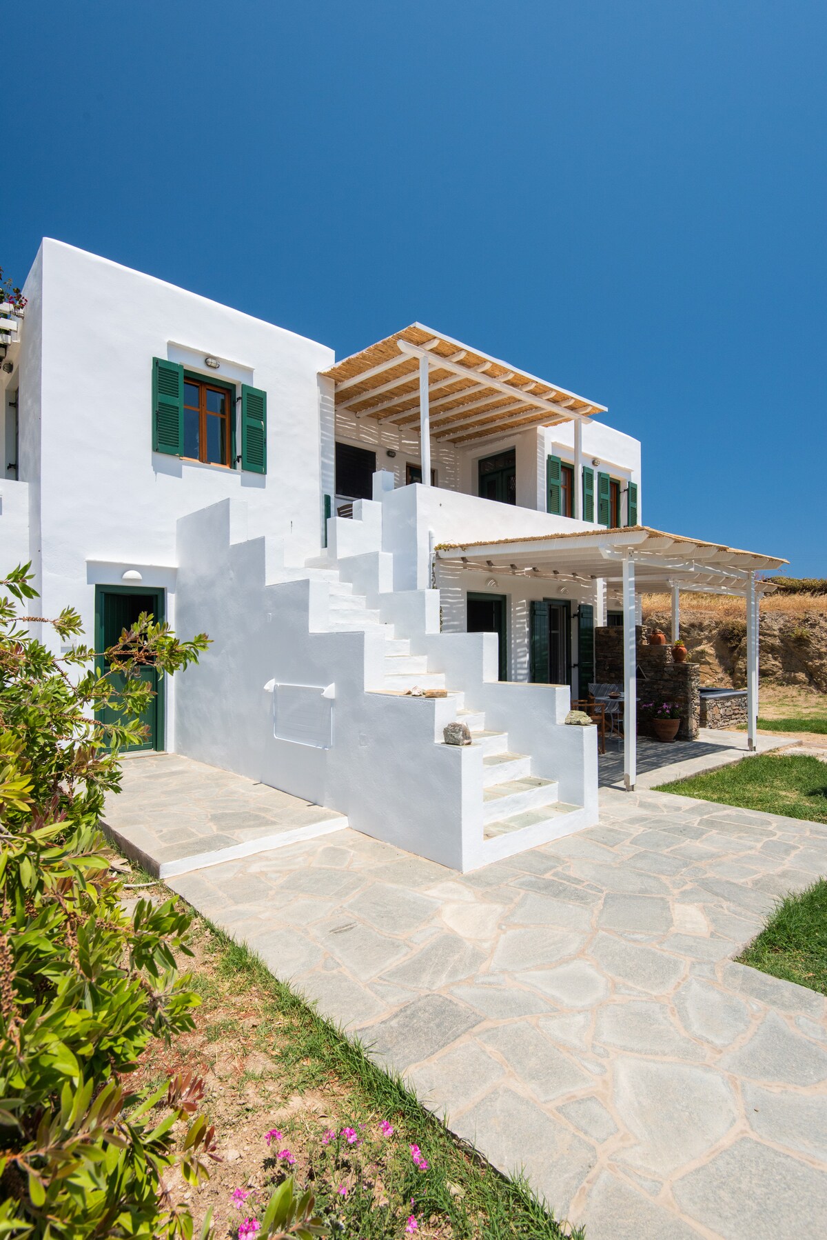 The exterior of a white building is seen, featuring green window shutters and a covered porch with a wooden trellis. Stone steps lead to the entrance, while a pathway lined with greenery and flowering plants adds to the welcoming atmosphere.