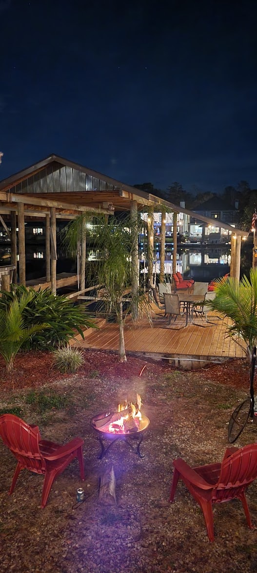 A cozy outdoor area is illuminated at night, featuring two red chairs positioned around a small fire pit. The deck is visible nearby, with tables and chairs arranged for gatherings. Reflections of light shimmer on the water, enhancing the tranquil ambiance.