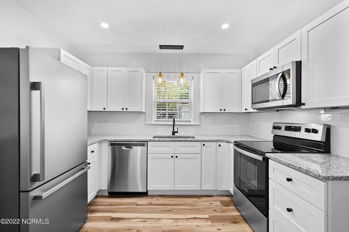 The modern kitchen features stainless steel appliances, including a refrigerator, dishwasher, and microwave. Bright white cabinets are complemented by a polished granite countertop and subtle lighting fixtures. A window above the sink allows natural light to enhance the space.