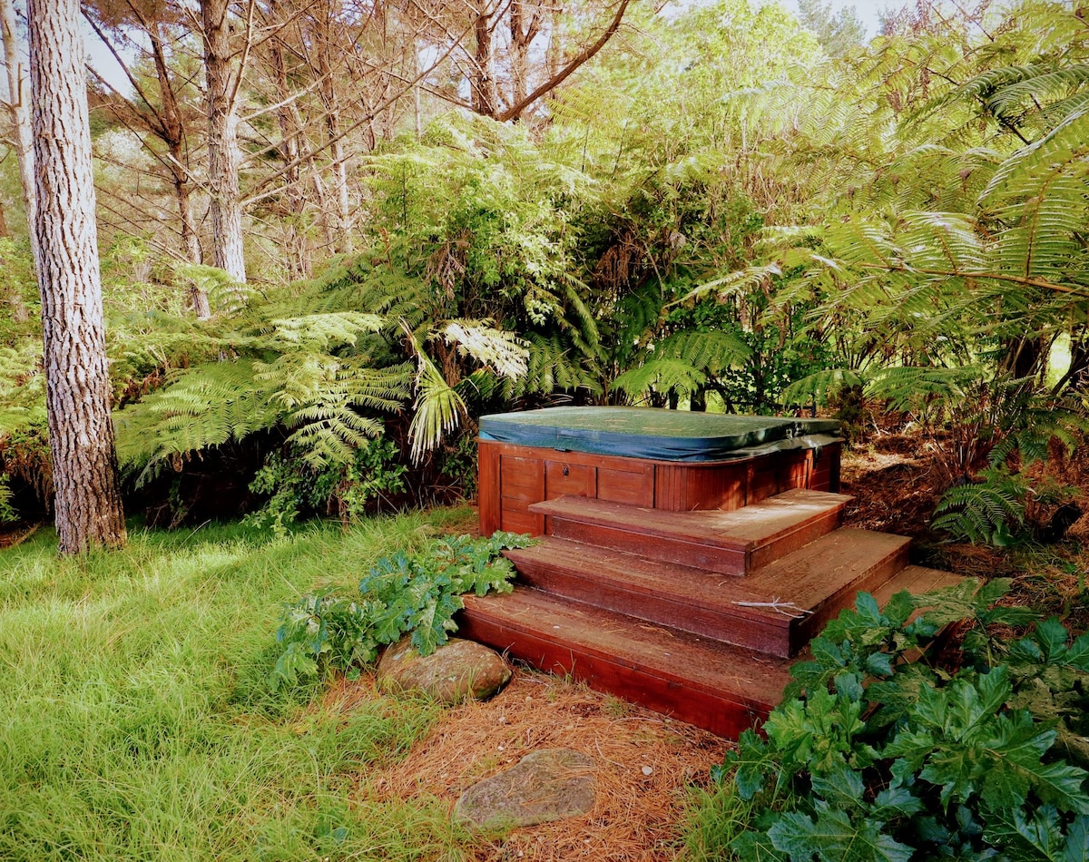 A wooden hot tub is nestled within a lush green setting, surrounded by tall trees and ferns. It is approached by a set of steps leading up from a grassy area, creating a secluded and tranquil experience in nature.