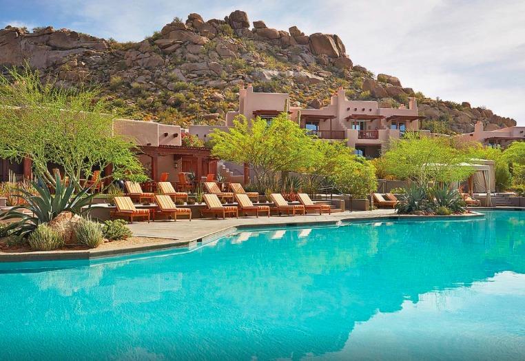 A sprawling swimming pool reflects bright blue skies, surrounded by natural desert landscaping. Comfortable lounge chairs are arranged along the pool's edge, offering serene views of the rocky hills in the background, dotted with greenery.