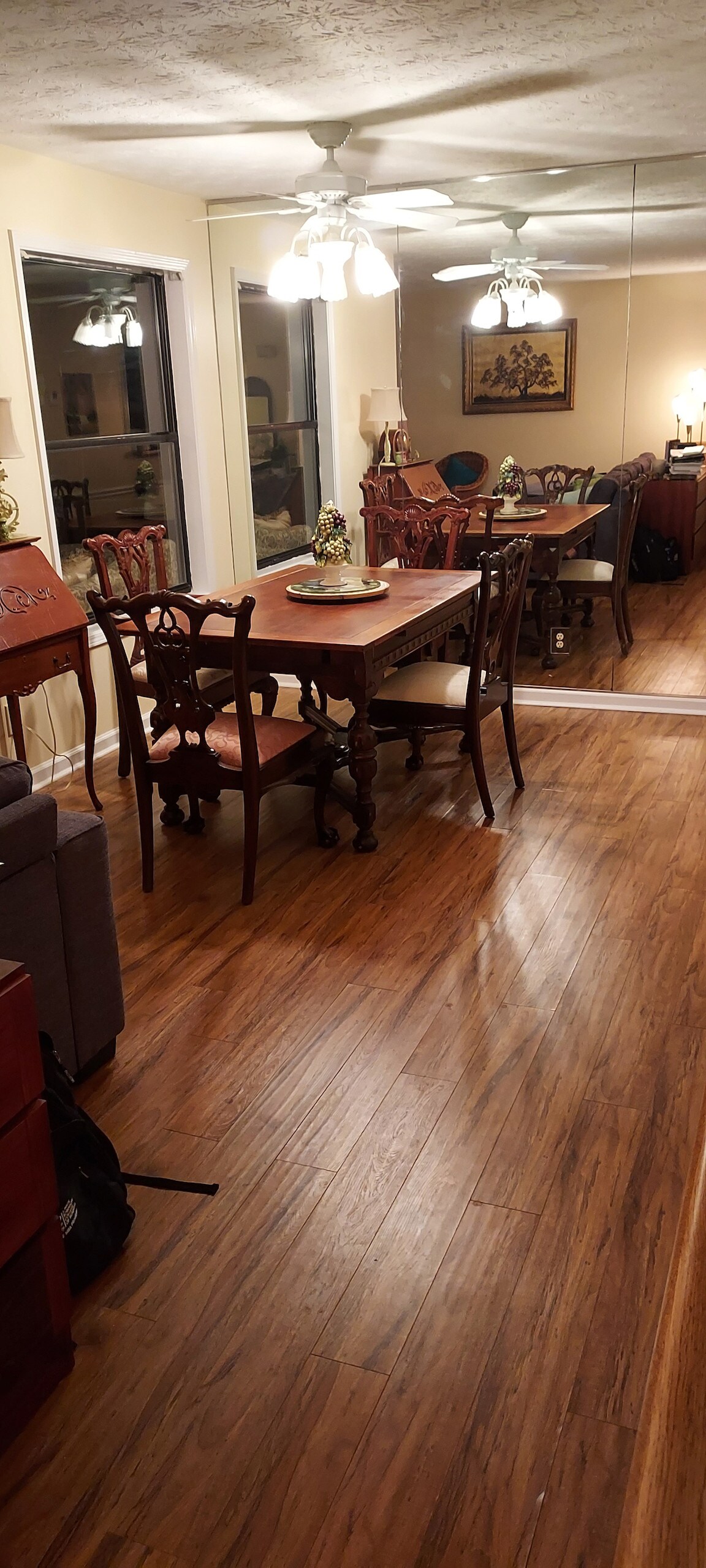 A wooden dining table is surrounded by elegantly designed chairs, set against a mirrored wall. Ceiling fans are positioned overhead, while a warm wood floor enhances the dining area. Windows allow natural light to fill the space, creating an open and airy feel.