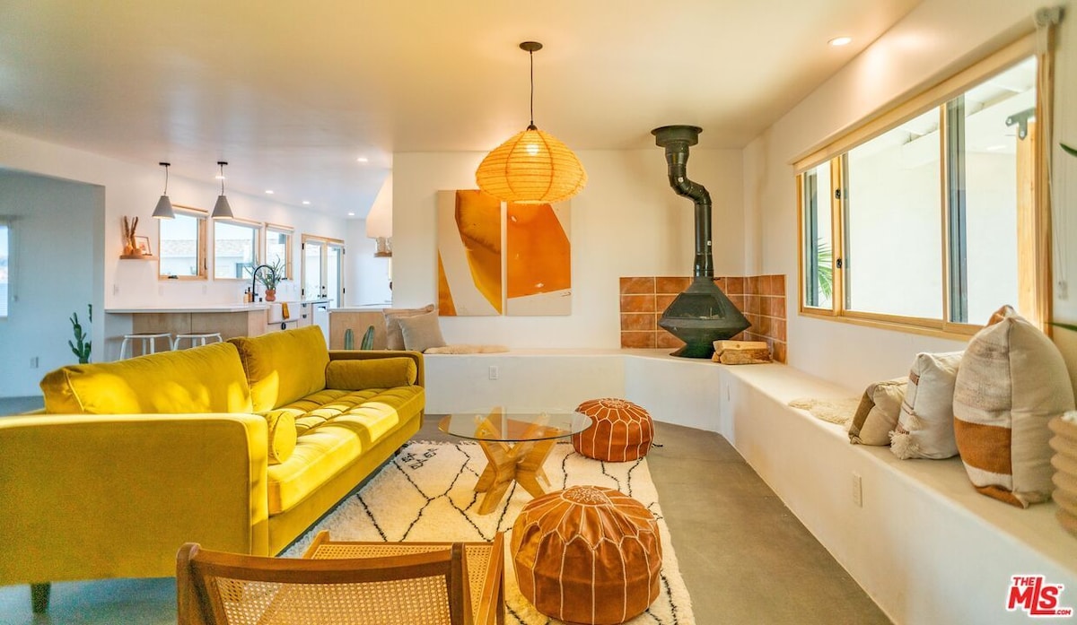A modern living area features a bright yellow sofa positioned across from a glass coffee table, with circular poufs nearby. Natural light floods through the large windows, illuminating the neutral-toned walls and polished concrete floors. A unique fireplace and decorative wall art enhance the space.