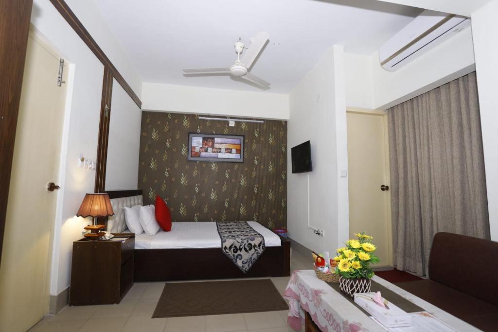 A comfortable room is presented with a spacious bed adorned with decorative pillows. A television is mounted on the wall opposite the bed. A small table with flowers adds a touch of color, while curtains and a ceiling fan enhance the ambiance.