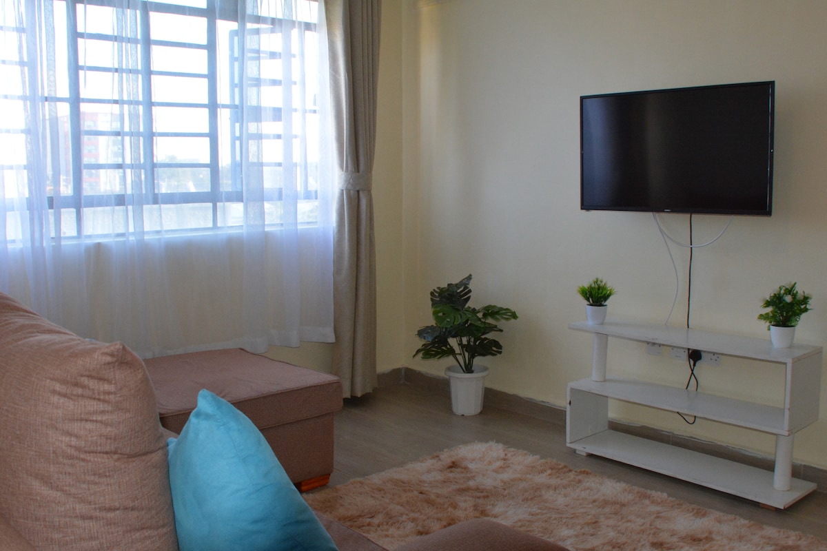 A cozy living area features a soft sofa with a light blue cushion and a plush area rug. Natural light filters through large windows dressed in sheer curtains. A wall-mounted television is positioned above a minimalistic white TV stand, complemented by potted plants.