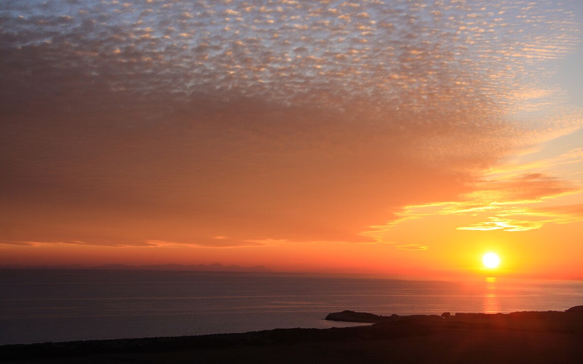 A sunset view over the ocean displays a gradient of warm colors, transitioning from orange to soft pink and purple. The sun sits low on the horizon, casting a gentle glow across the water, while the sky is adorned with textured clouds.