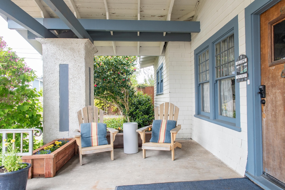 A welcoming porch area is presented with two wooden Adirondack chairs adorned with pillows, complemented by a small side table. Lush greenery and flowering shrubs create an inviting outdoor space, with natural light filtering through the open architecture of the roof.