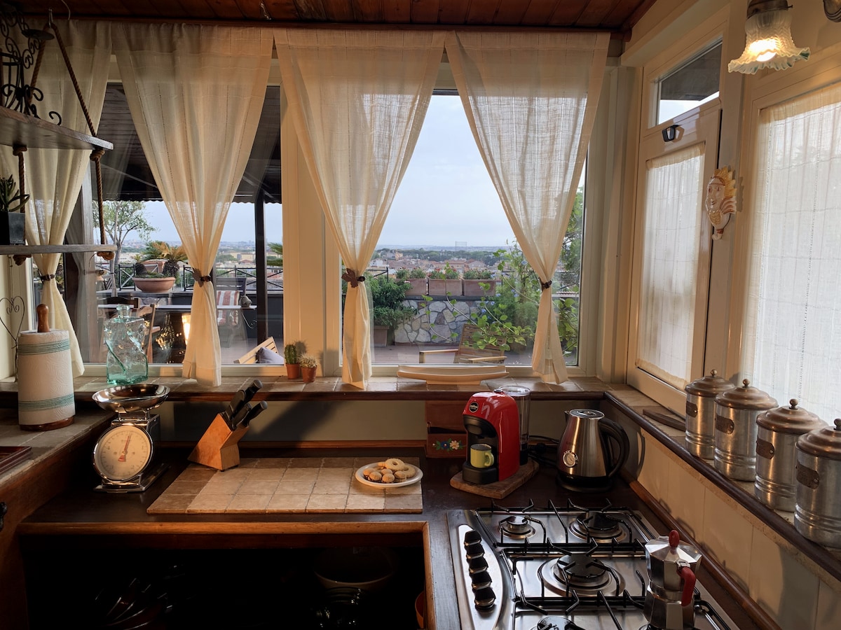 A rustic kitchen features wooden countertops and modern appliances. Light filters through sheer curtains, framing a view of rooftops and greenery. A tray of cookies sits on the counter next to a kettle and a red coffee maker, creating a cozy cooking space.