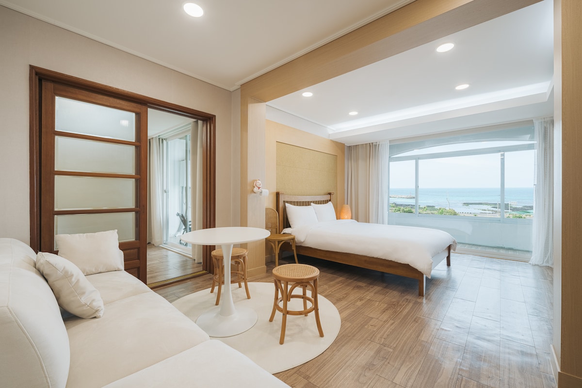 A bright and airy room features large windows offering sea views. A spacious bed is accompanied by a small round table and chairs. A comfortable sofa sits opposite, enhancing the room's relaxation options. Natural light fills the space, accentuated by neutral-colored walls.