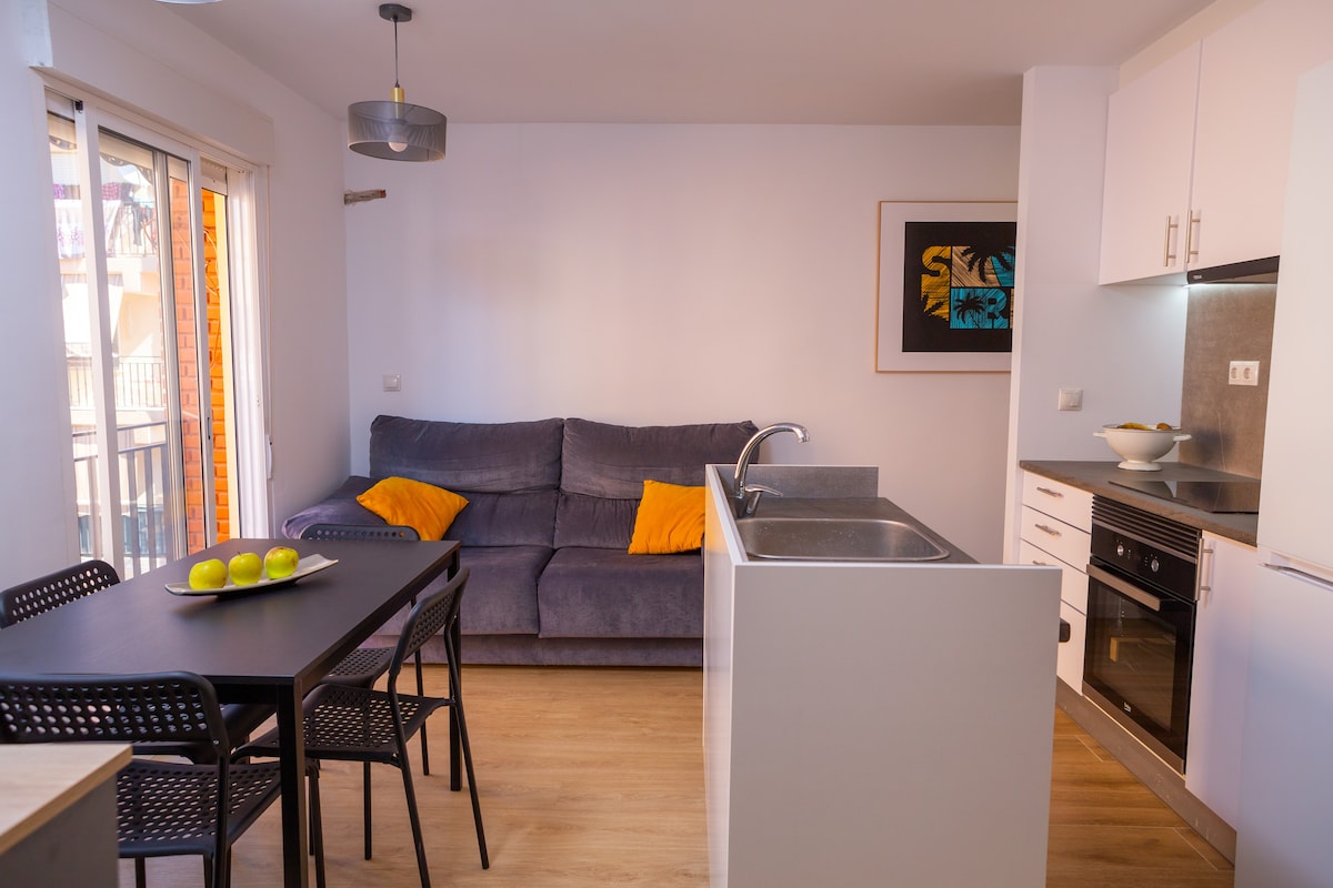 A modern living area features a comfortable gray sofa adorned with orange cushions. A dining table with four black chairs is positioned nearby. The kitchen area includes sleek cabinets and a stainless-steel sink, with light streaming in through glass doors that lead to a balcony.