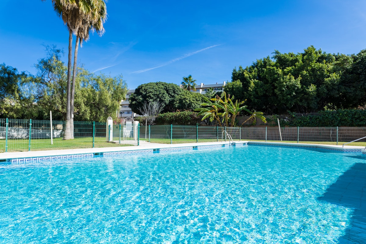 A community swimming pool is depicted, featuring clear blue water under bright sunlight. Lush greenery surrounds the area, with palm trees in the background. A fenced perimeter ensures safety, and lounge chairs are arranged nearby for relaxation.