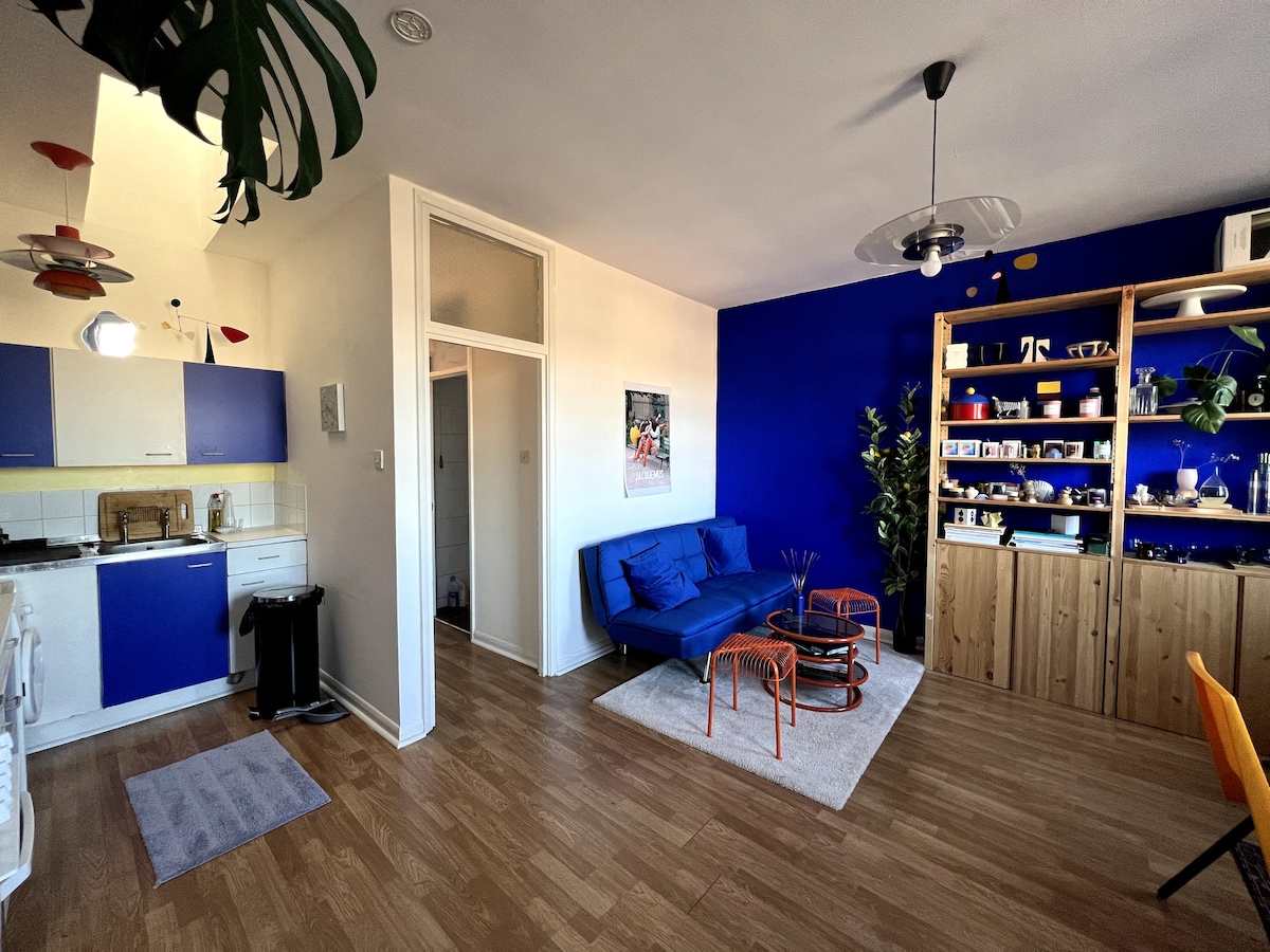 A bright living area features a blue sofa complemented by orange accent tables. The kitchen is equipped with blue and white cabinetry. Shelving units display decorative items and plants. Natural light fills the space through large windows, enhancing the cheerful atmosphere.