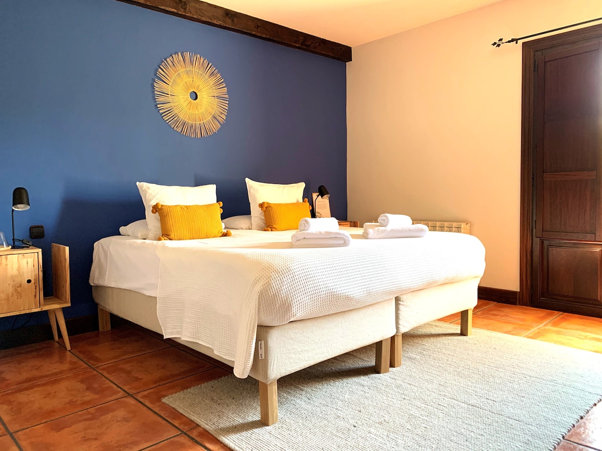 A spacious bedroom features two beds with light bedding, accented by yellow decorative pillows. A textured area rug covers the tiled floor, and a round wall decoration adds character. Natural light enters through a wooden window, enhancing the calming atmosphere.