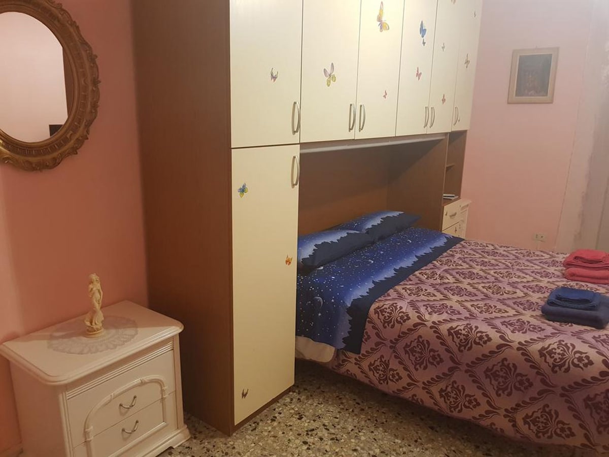 A bedroom features a spacious bed covered in a patterned blanket and stacked towels on top. A nightstand with a decorative statue is positioned beside the bed. The walls are painted pink, with whimsical butterfly designs on the cabinetry and artwork in the background.