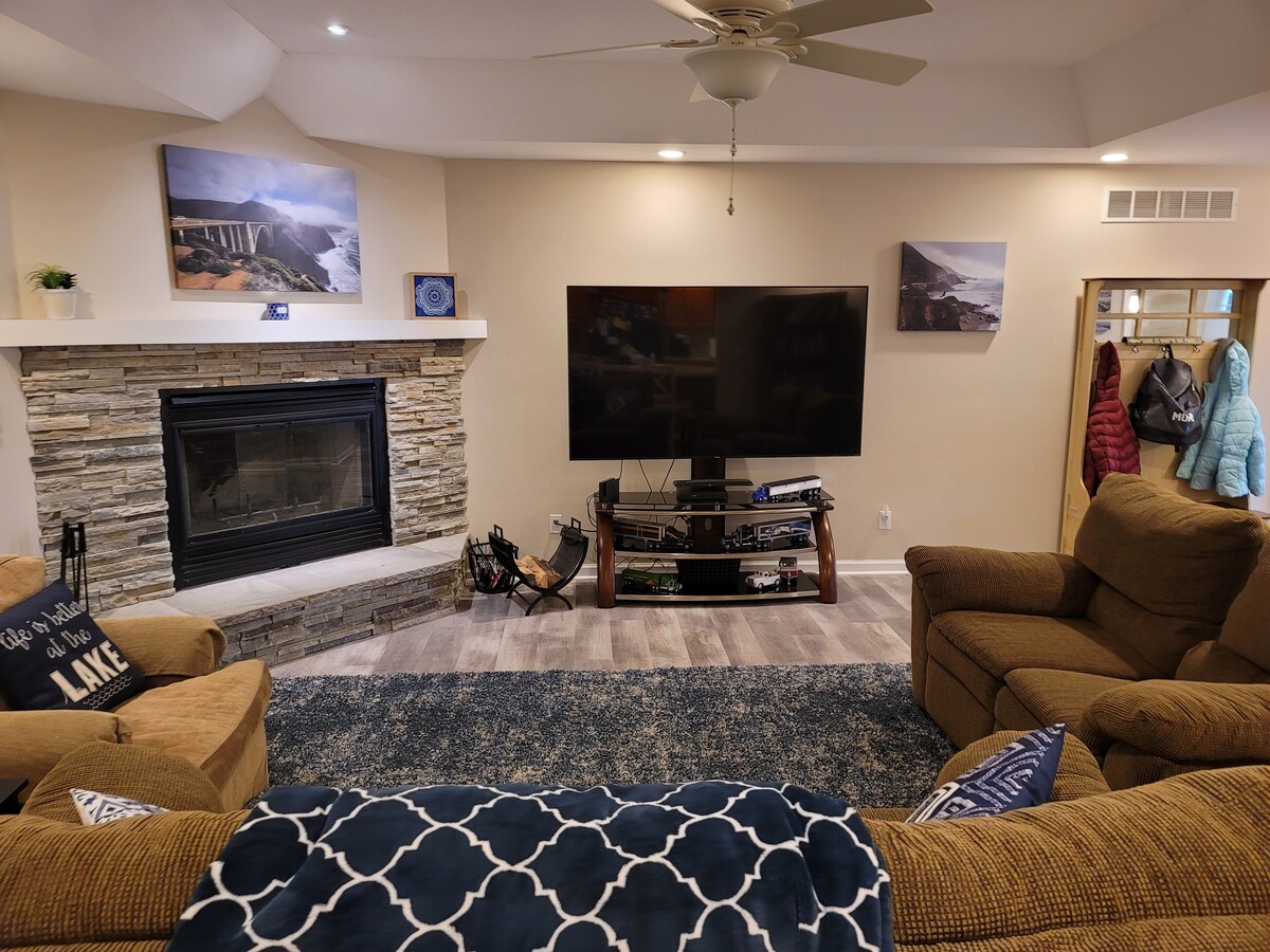 A cozy living area features a stone fireplace with a mounted television above it. Two inviting sofas face the fireplace, accompanied by decorative pillows. Natural light brightens the space, which is accented by framed landscape artwork on the walls and a plush area rug on the floor.