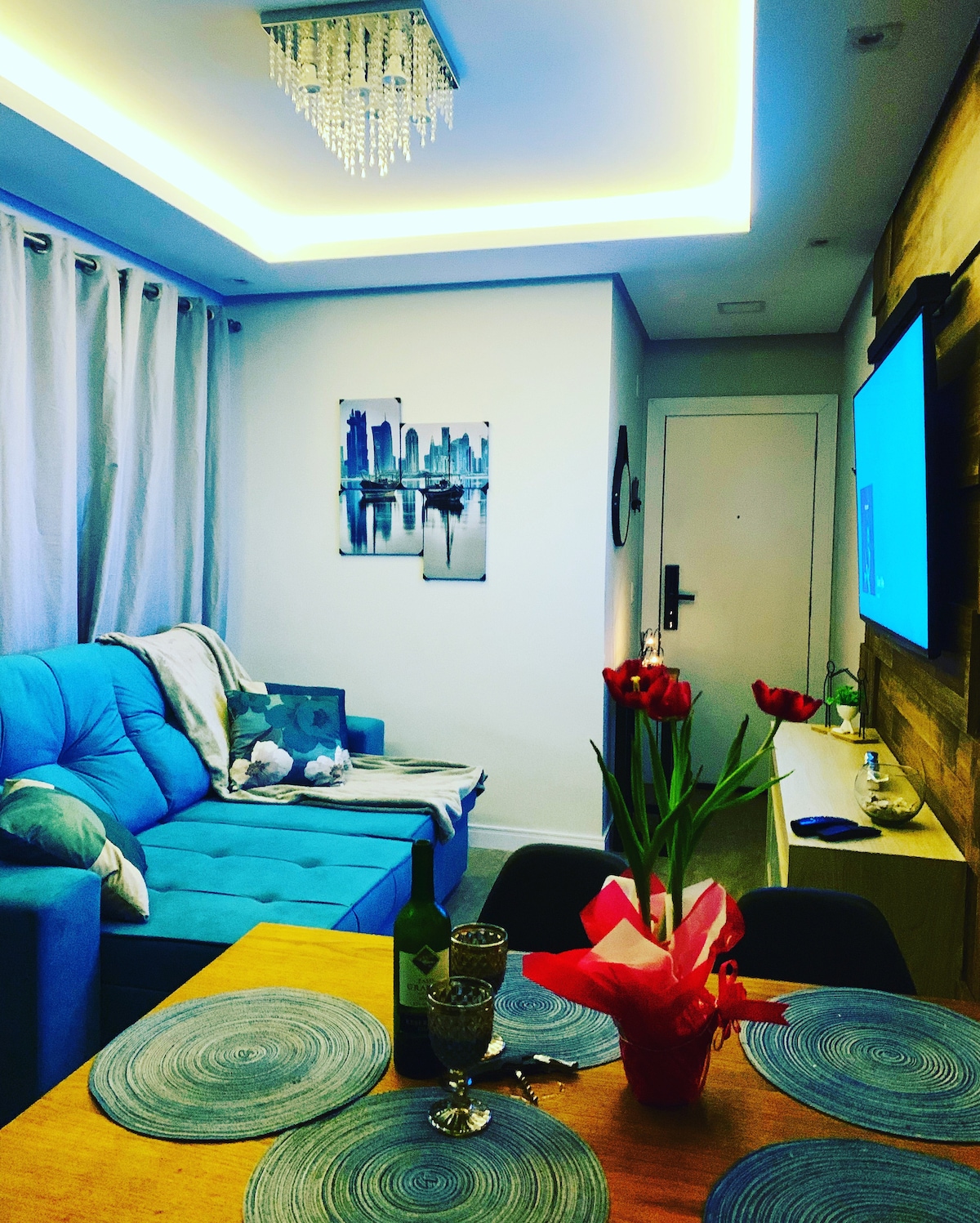 A well-lit living area features a blue sofa with decorative pillows, complemented by a small dining table set with two glasses and a flower centerpiece. A modern art piece depicting a city skyline is hung on the wall, and a TV is mounted opposite the seating area.