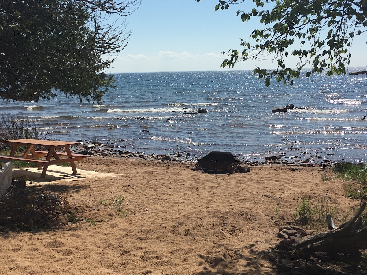 A sandy beach area leads to the shimmering waters of Lake Superior, visible under a clear blue sky. A wooden picnic table is positioned on the sand, offering a space for outdoor dining. Smooth stones and a few trees frame the scene, enhancing the natural setting.