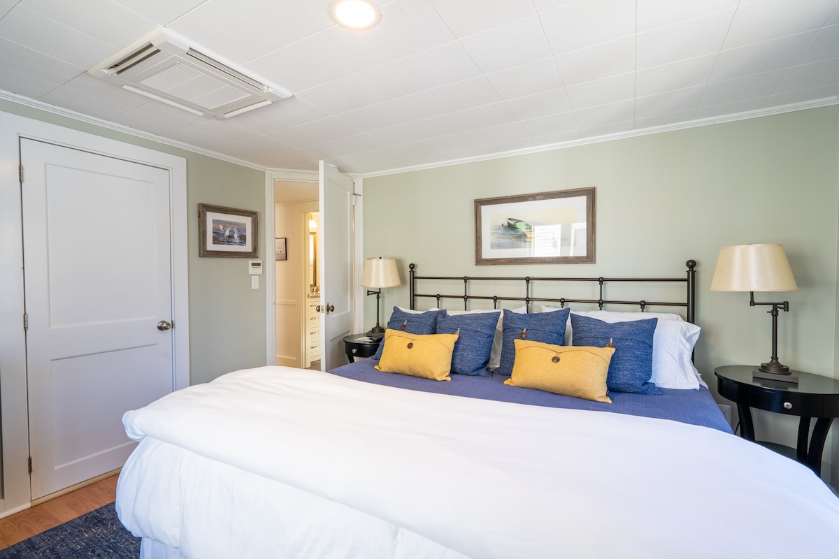 A cozy bedroom is presented with a spacious bed dressed in crisp white linens and a navy blue blanket. Yellow accent pillows add a pop of color. A bedside table is placed on either side, while soft lighting from the ceiling enhances the room's overall comfort.
