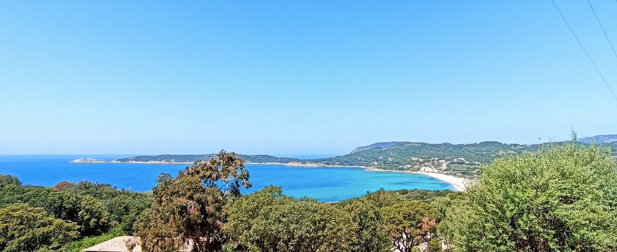 A panoramic view of the coastline features clear blue waters and a sandy beach, framed by lush green foliage. The landscape includes distant hills and the serene sea, under a bright blue sky, creating a sense of tranquility.