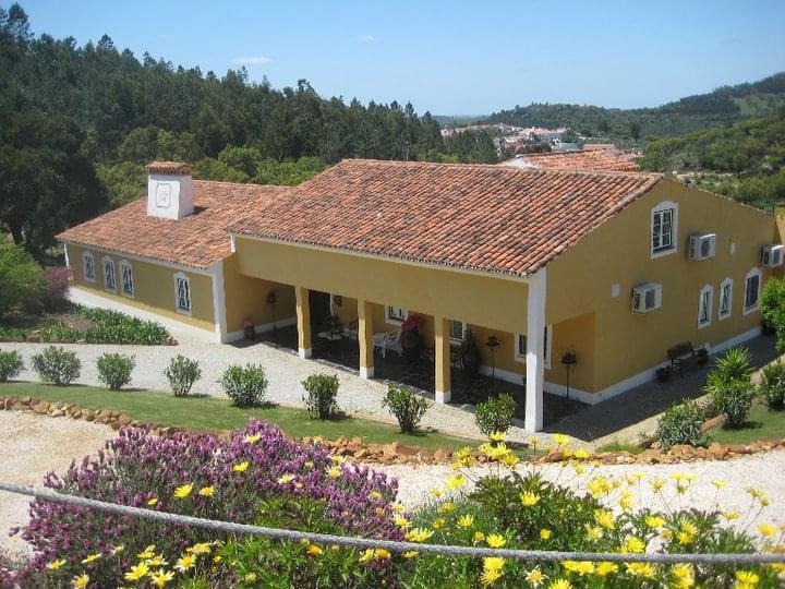 A charming yellow house with a red-tiled roof is surrounded by lush greenery and colorful flower beds. Multiple windows are visible, allowing for plenty of natural light. The inviting outdoor area includes a well-maintained garden and a gravel pathway leading to the entrance.
