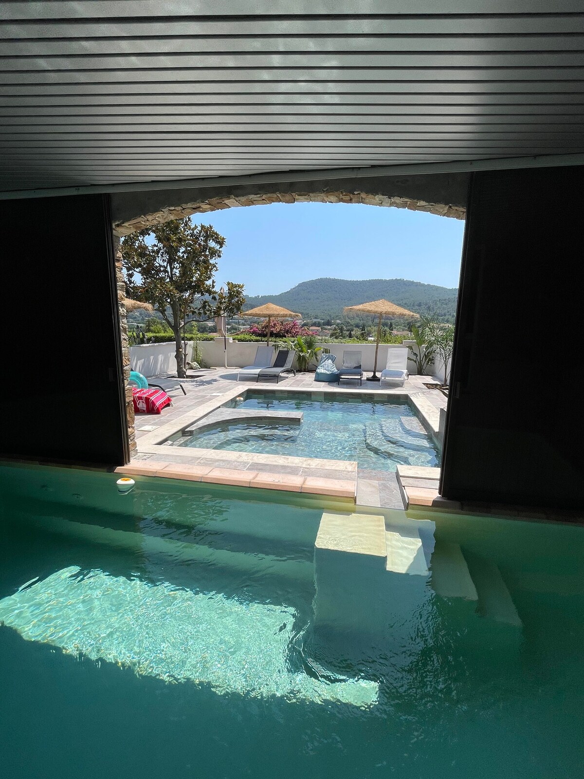 An outdoor pool area is visible, featuring a spacious pool surrounded by lounge chairs and umbrellas. The scene is framed by arches, with hills and trees in the background, providing a serene view and an inviting outdoor space.