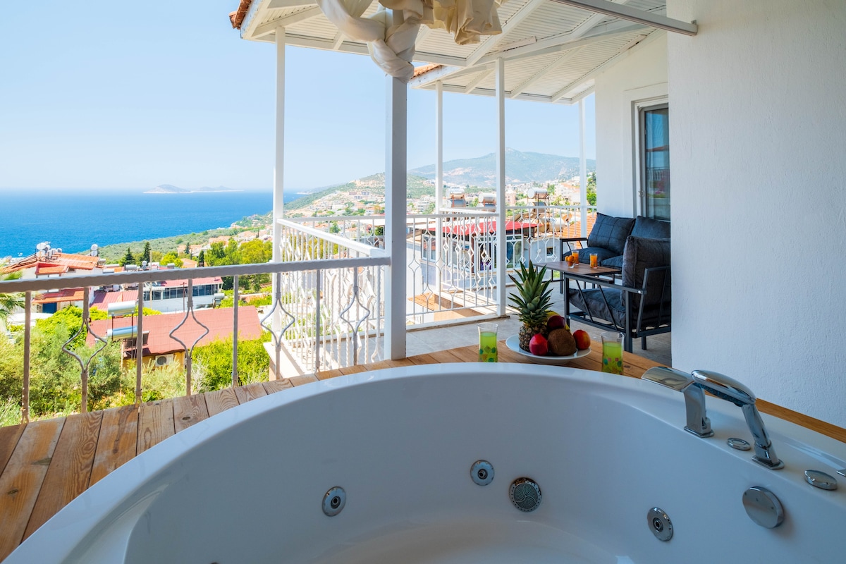 A jetted jacuzzi is positioned on a wooden deck, offering views of the ocean and surrounding landscape. The terrace includes comfortable seating arrangements with a small table, providing an inviting space to relax and enjoy scenic vistas.