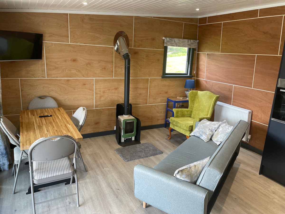 An inviting living area features a comfortable sofa and a wooden dining table. A small green stove is positioned near the wall, with a blue side table and a lamp nearby. The walls are adorned with warm wood paneling, creating a cozy environment.