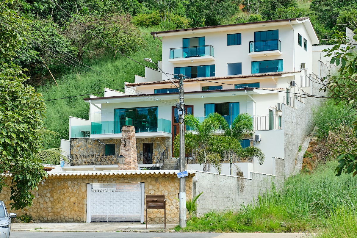 The exterior of the three-story house is showcased, featuring a combination of stone and modern materials. Large windows offer views of the surrounding greenery, while balconies provide outdoor space. The entrance is enhanced by palm trees, contributing to the home's inviting appearance.