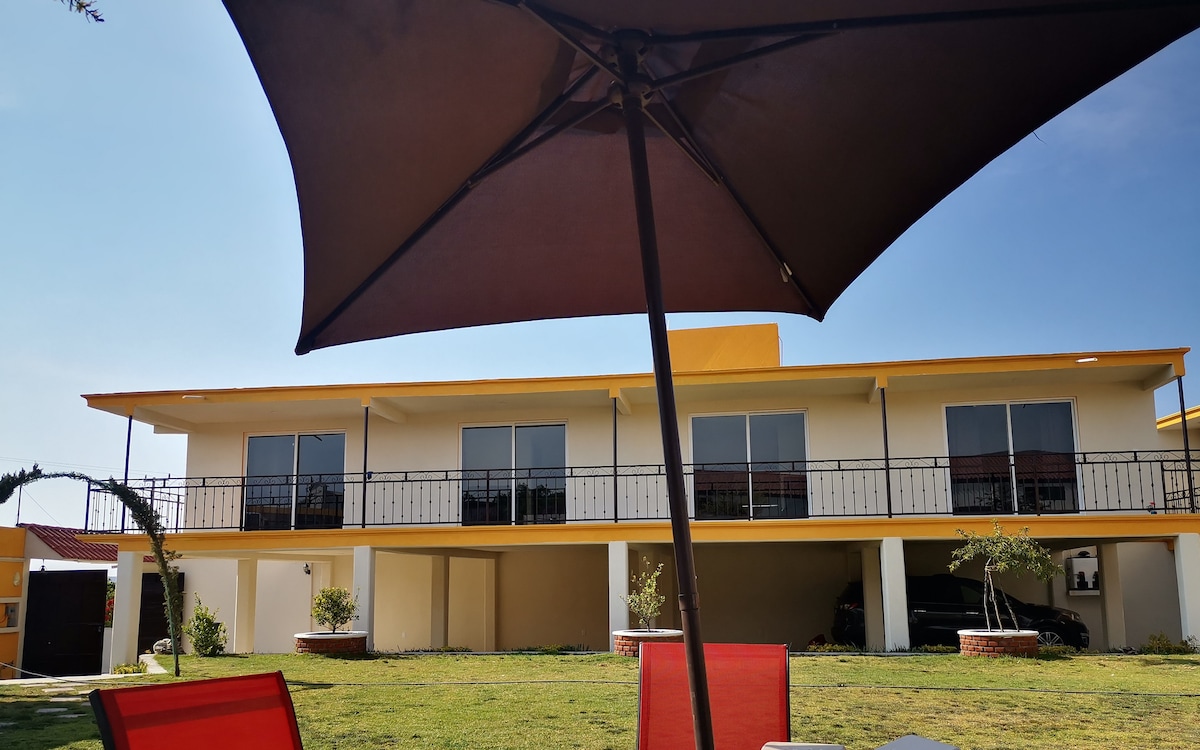 The image displays a two-story villa with a yellow exterior and balconies, framed by a grassy area. Sunlight reflects off the large windows, while chairs and an umbrella are positioned in the foreground, providing shade and a relaxed seating option.