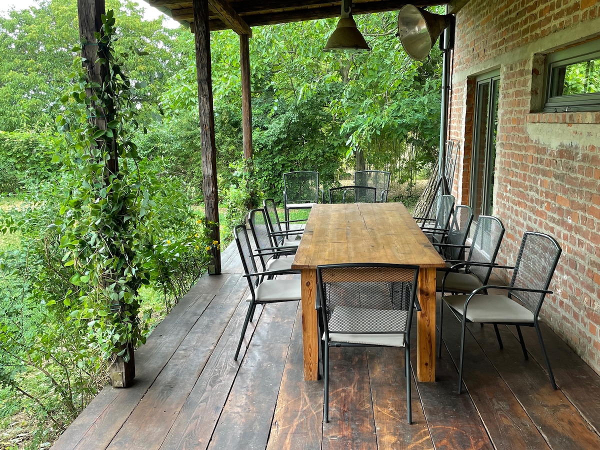 An outdoor dining area features a large wooden table surrounded by multiple metal chairs. Lush greenery frames the space, and wooden beams support a covered porch. Natural light highlights the rustic brick walls, creating a serene environment for gatherings.