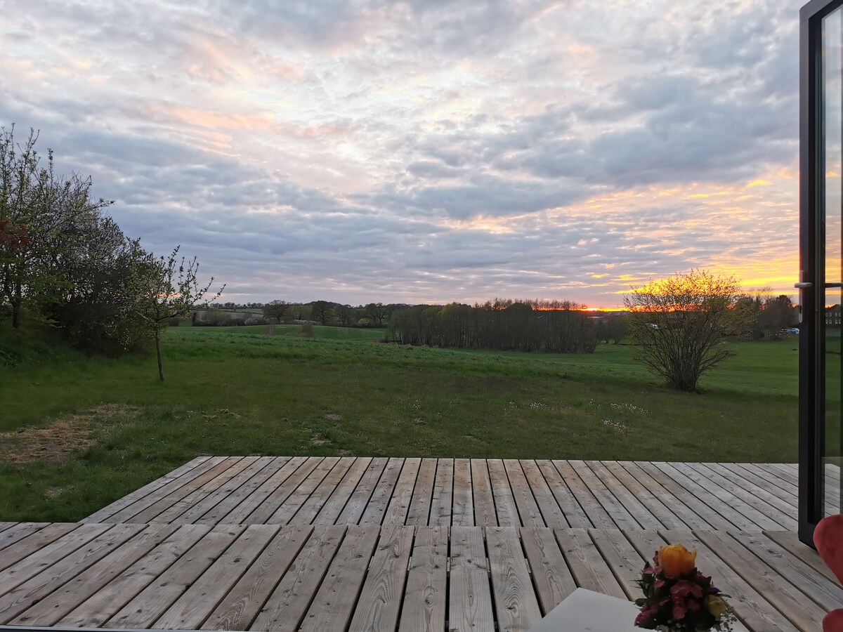 A wooden terrace opens up to a vast green landscape, with soft grass and scattered trees in the background. A colorful flower arrangement is placed on the terrace. A serene sky filled with clouds reflects warm hues of orange and pink as the sun sets.