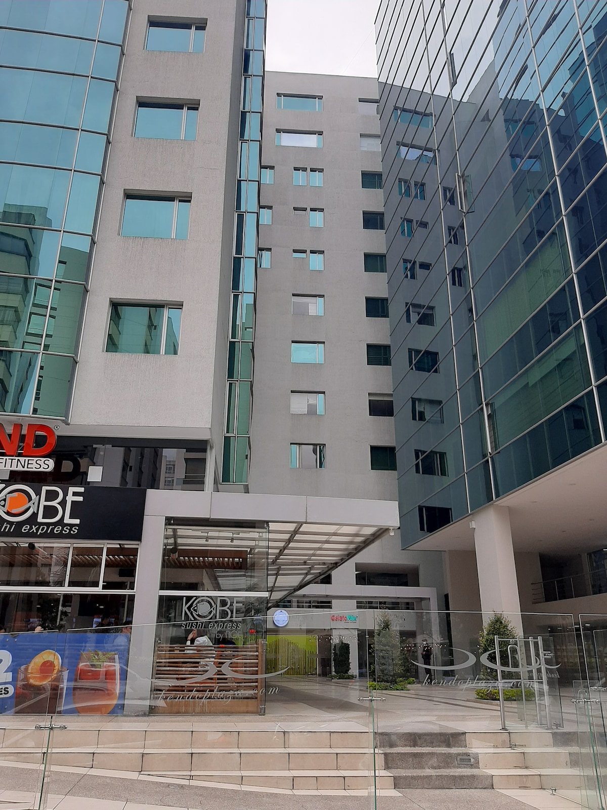 An exterior view of a modern multi-story building features large glass windows reflecting the surrounding environment. The entrance is marked by a covered walkway with a glass canopy. Nearby, a fitness center sign can be seen, adding to the commercial aspect of the area.