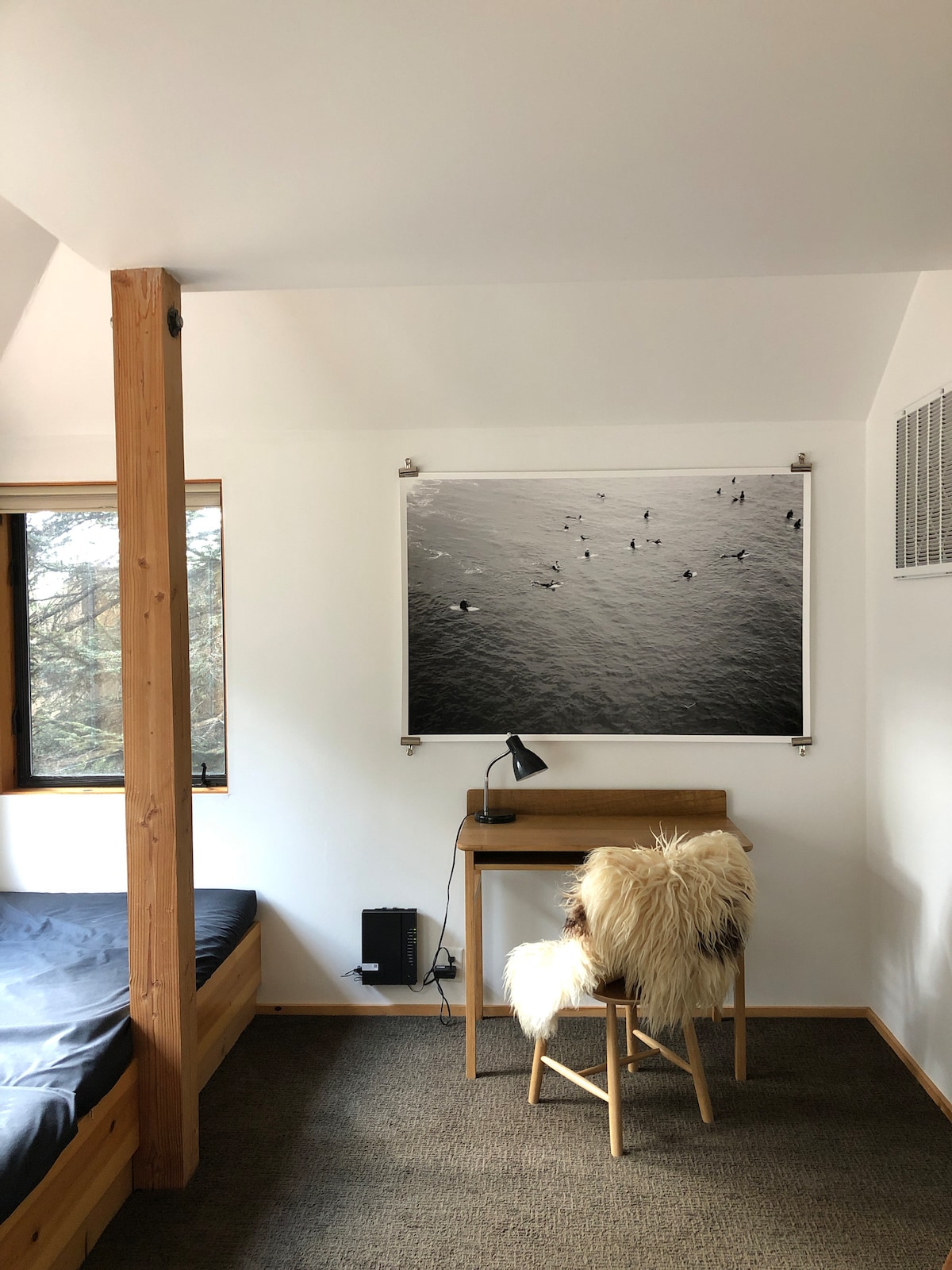 A serene bedroom is illustrated, showcasing a spacious bed with muted bedding. A wooden desk and chair, adorned with a cozy faux fur throw, are positioned by a window. A striking black-and-white photograph of surfers on water hangs on the wall, enhancing the calming ambiance.