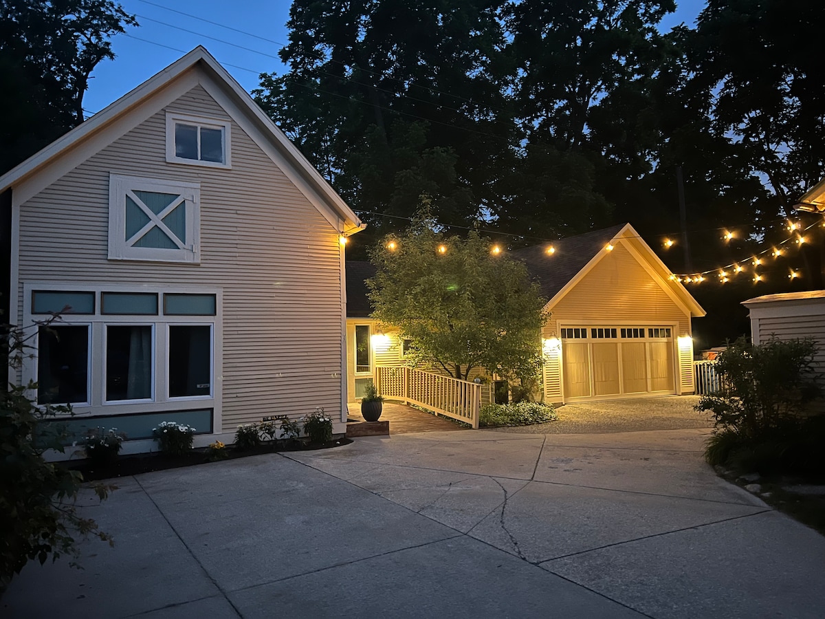 The exterior of the Carriage House is illuminated by warm string lights, creating a welcoming ambiance at dusk. The house features large windows, a decorative front porch, and a spacious driveway that leads to a garage. Lush greenery surrounds the property.