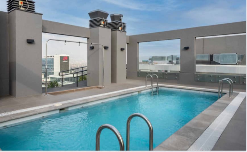 A rooftop pool area features clear, inviting water surrounded by sleek grey walls and modern architectural elements. Handrails lead into the pool, and large windows provide expansive views of the city skyline, creating a bright and open atmosphere.