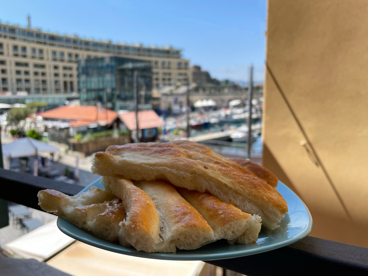 A plate of golden-brown breadsticks is displayed on a balcony railing. The background features a view of a bustling marina with boats and waterfront buildings under a clear blue sky.