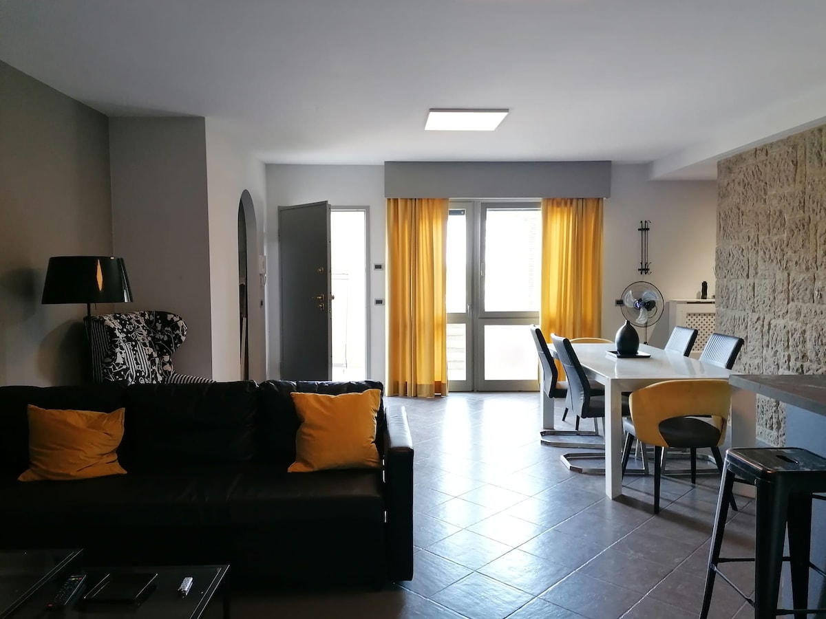 A modern living area features a dark sofa adorned with yellow cushions. A dining table with six chairs is illuminated by natural light from large windows, dressed in bright yellow curtains. A decorative fan and stylish lighting elements add to the space's contemporary design.