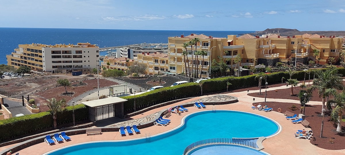 A panoramic view shows a large swimming pool surrounded by tropical gardens and palm trees. Colorful sun loungers are arranged along the poolside. In the distance, the Atlantic Ocean meets the horizon, with residential buildings situated nearby in a tranquil setting.