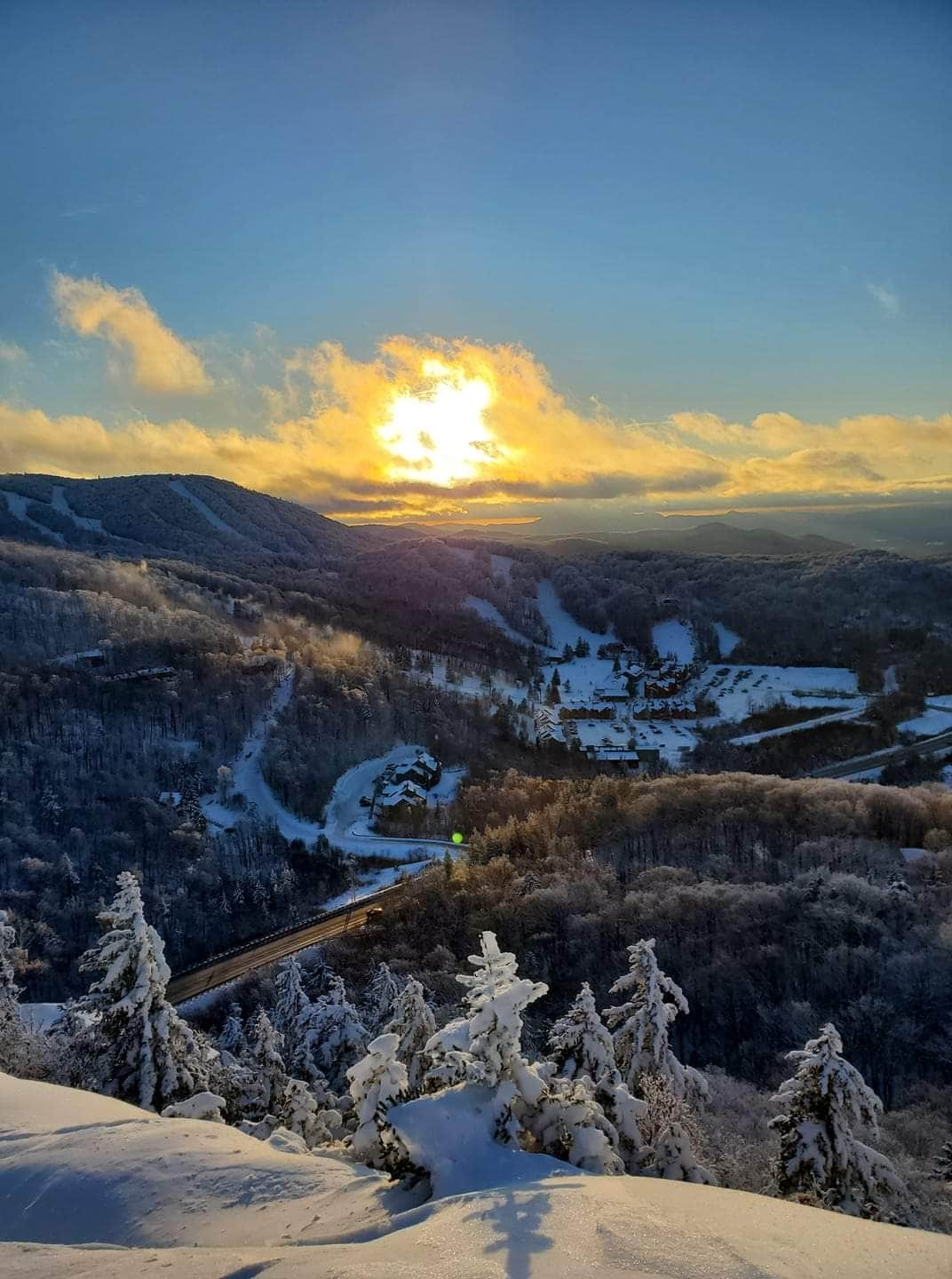A stunning winter landscape is captured featuring snow-covered mountains and a brilliant sunset. A valley is visible with a winding road, and trees covered in fresh snow add to the serene atmosphere, creating a breathtaking view of the natural surroundings.