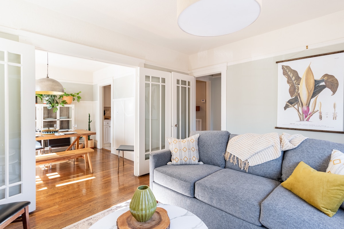 A bright living room features a comfortable blue sofa adorned with patterned cushions. Nearby, a dining area with a wooden table is partially visible through glass-paned doors. Natural light enhances the space, while a framed botanical print hangs on the wall.