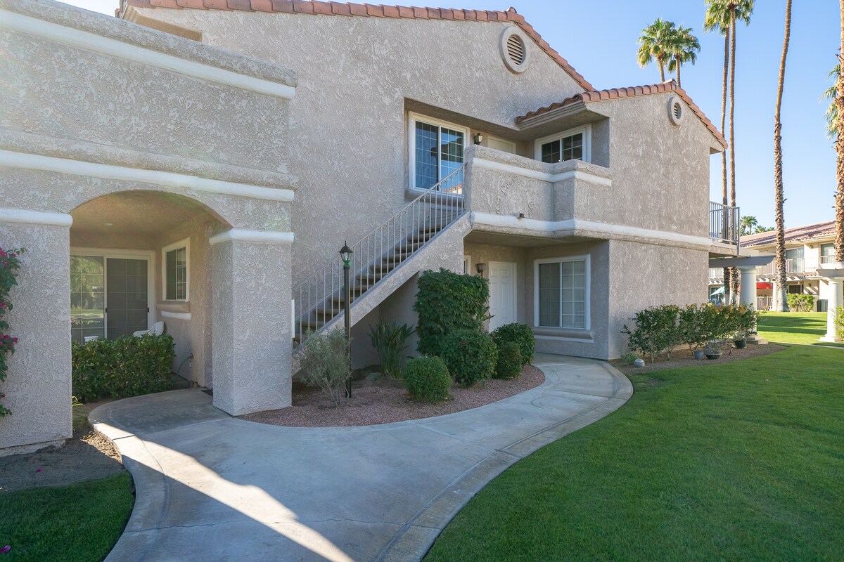 The exterior of the condo features a light-colored stucco facade with a tiled roof. Landscaping includes manicured grass and small shrubs. A pathway leads to the entrance, while a set of stairs ascends to the upper level, flanked by palm trees and well-maintained grounds.