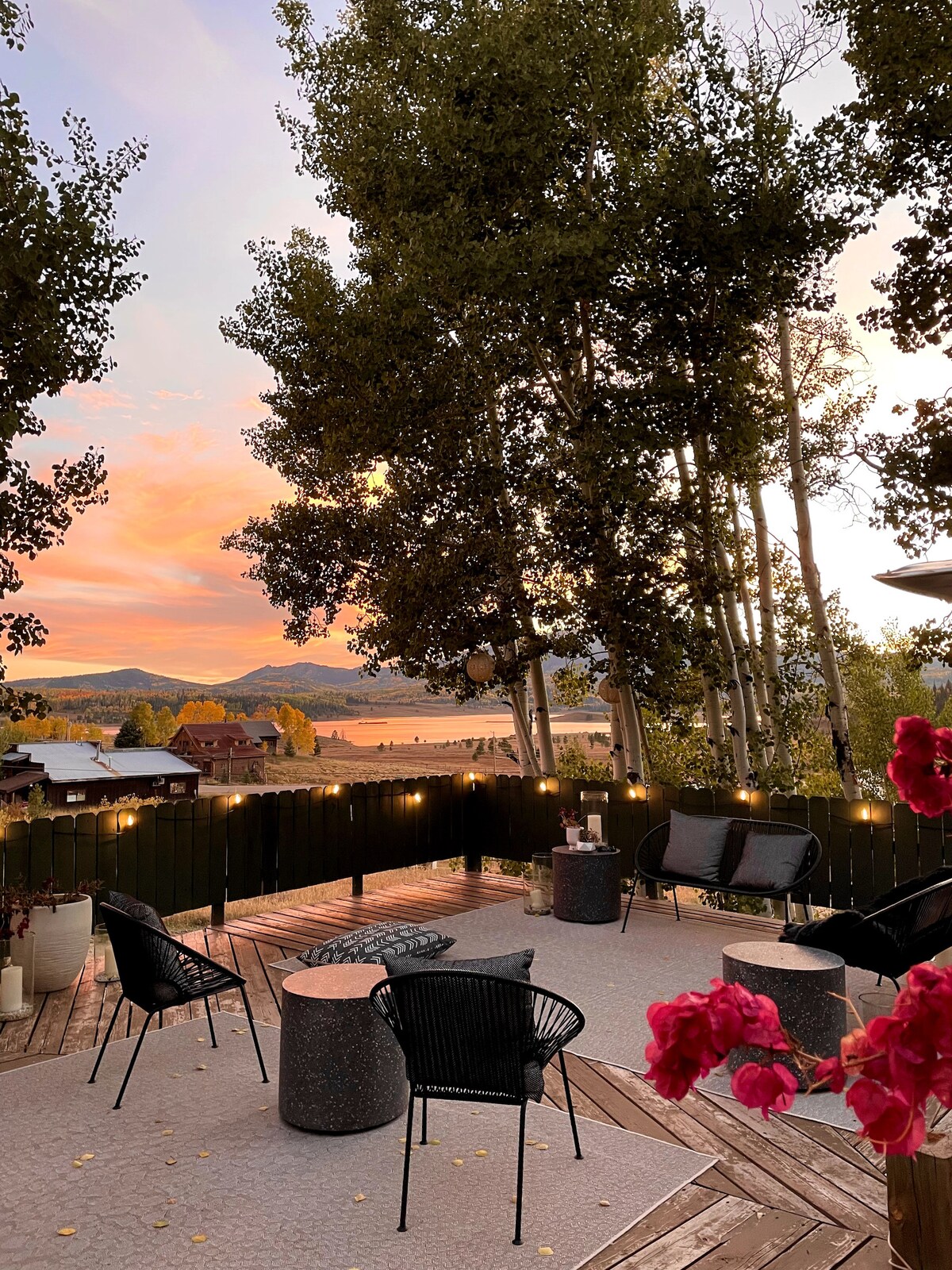 An outdoor deck features black wicker seating arrangements and small side tables surrounded by trees. Soft lighting illuminates the space, highlighting a view of the lake and distant mountains, showcasing a colorful sunset in the sky.