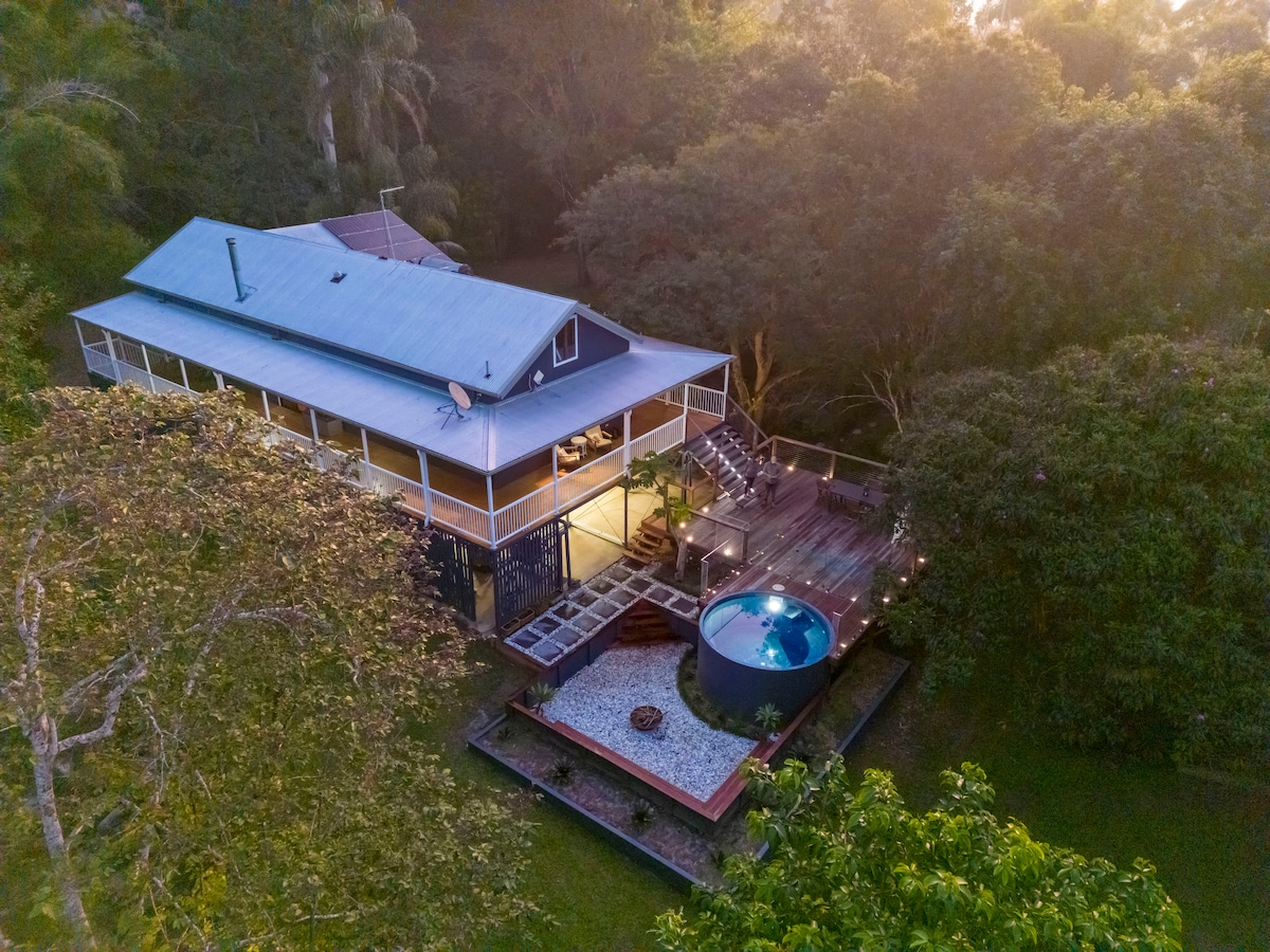 A modern Queenslander home is depicted from above, surrounded by lush greenery. A magnesium plunge pool is situated near a wooden deck, with stairs leading up to the entrance. The area is softly illuminated by warm lighting, creating a serene setting within the natural landscape.
