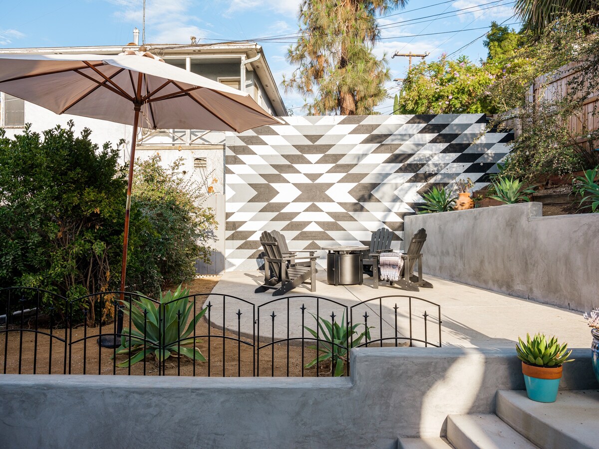 An outdoor seating area is showcased, featuring several Adirondack chairs arranged around a central fire pit. A large umbrella provides shade, while decorative plants and a patterned wall add visual interest. The space is designed for relaxation and social gatherings.