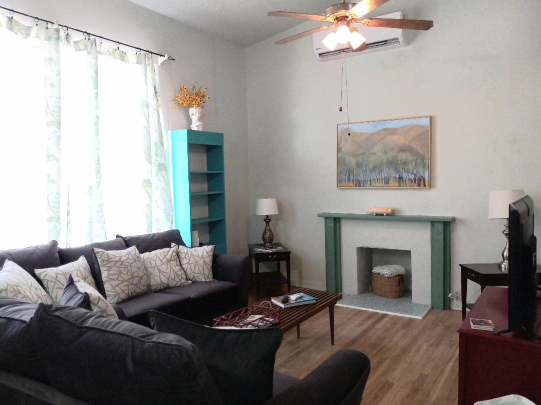 A comfortable living area features a dark sectional sofa adorned with decorative pillows. A wooden coffee table is positioned in front, complemented by an inviting art piece above the fireplace. Natural light enters through large windows with sheer curtains, enhancing the room's spaciousness.