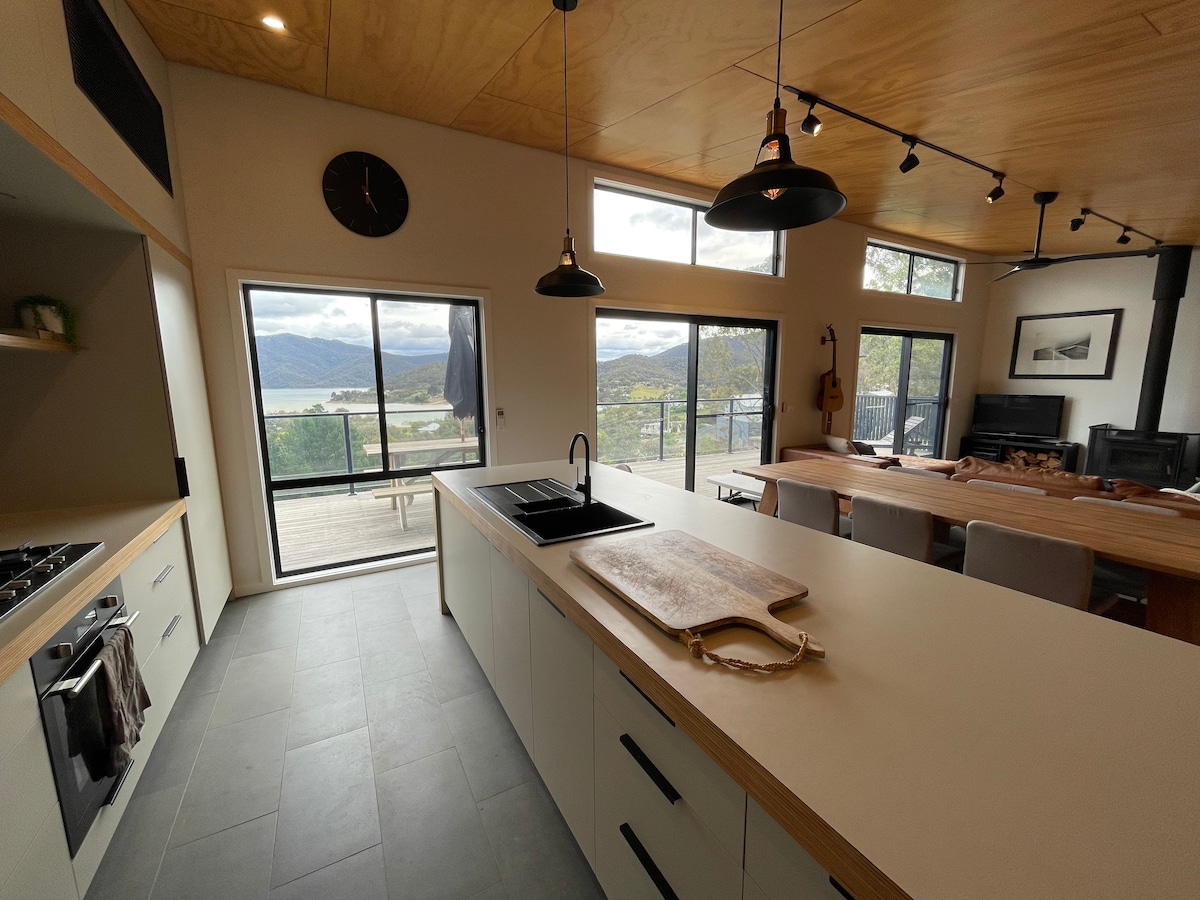 A modern kitchen features a centrally placed island with a sleek black sink. Large glass doors frame views of the lake and mountains beyond, while wooden accents are present in the ceiling and dining area, enhancing the open-plan layout.