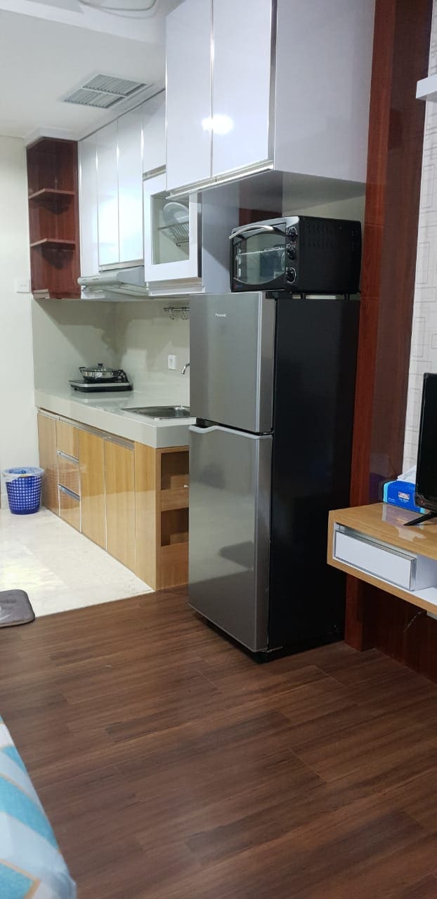 A compact kitchen area is visible, featuring sleek cabinets and a countertop. A stainless steel refrigerator stands beside a black microwave, while a small stove is placed against the wall. The floor showcases a warm wood finish, contributing to a functional yet inviting space.