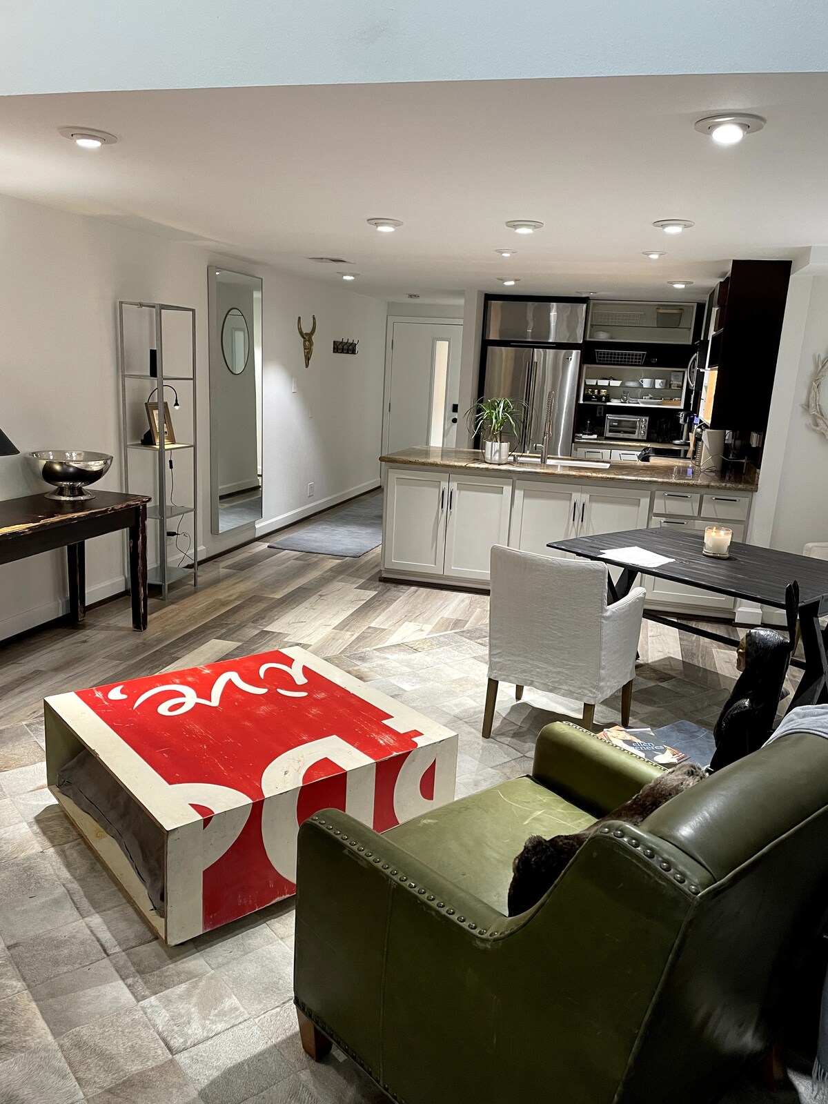 An open living space features wood-grain vinyl flooring and well-lit areas. A unique coffee table, designed with the Coca-Cola logo, sits in front of a leather reading chair. The kitchen includes modern appliances, and a dining table for four enhances the functional layout.