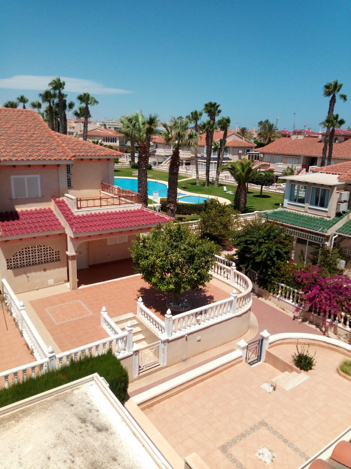 A view from above features a spacious property with a lower-level terrace leading to landscaped gardens. Palm trees are seen throughout the area, and a swimming pool is visible in the background, surrounded by additional residences.