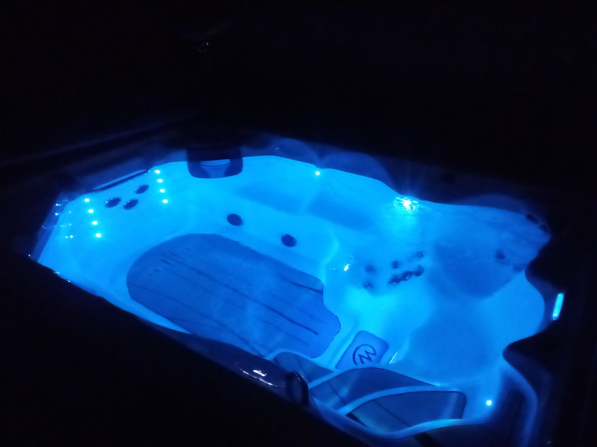 A hot tub is illuminated with soft blue lighting, creating a calming atmosphere. The sleek design includes built-in seating and jets, inviting relaxation. The surrounding area is dark, emphasizing the tranquil glow of the water.